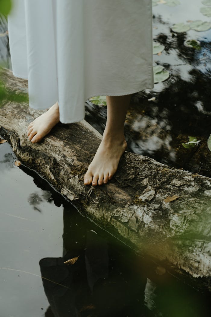 Barefoot woman stands on a log by a pond, surrounded by nature in a serene setting.